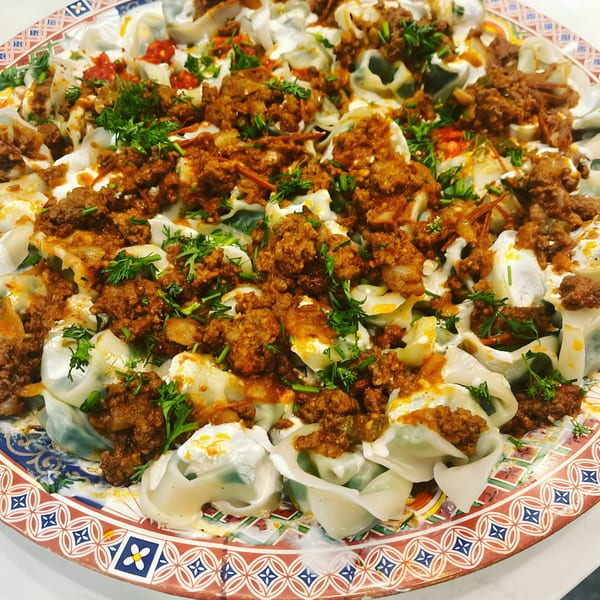 A large platter of Afghan Mantu dumplings, topped with ground meat sauce, yogurt, and fresh herbs.