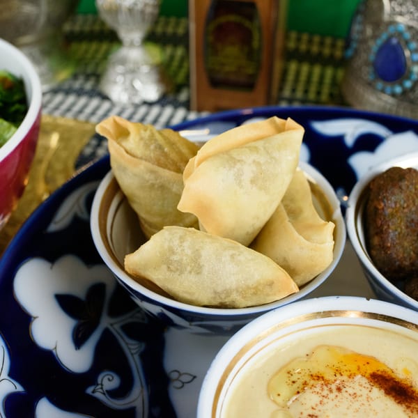 Close-up of golden fried Afghan samosas, hummus, and falafel on a decorative blue and white plate.